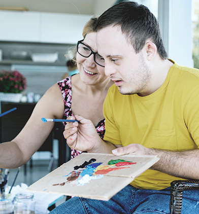 A smiling woman and a focused man engaging in a fun painting activity together at a table, with the woman guiding the man's hand holding a paintbrush over a colorful palette.